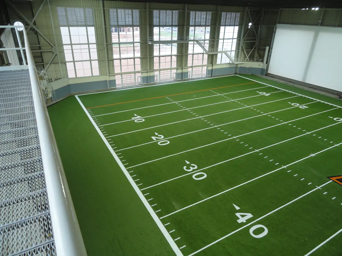 Indoor football field with vibrant green turf and white yard lines, viewed from an elevated metal walkway. Large windows provide natural light.