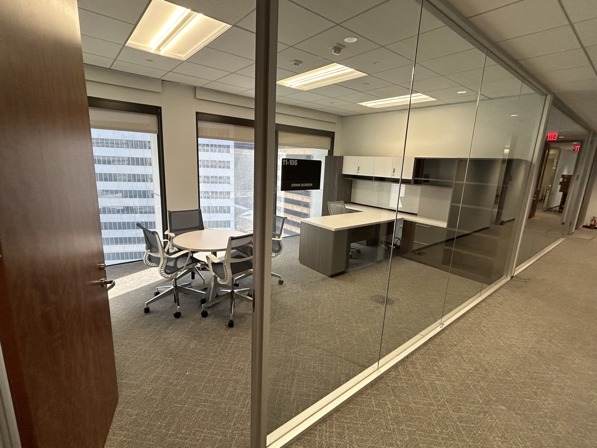 Modern office with glass walls, a round table with four chairs, and a desk. Large windows reveal a cityscape. Bright, professional atmosphere.