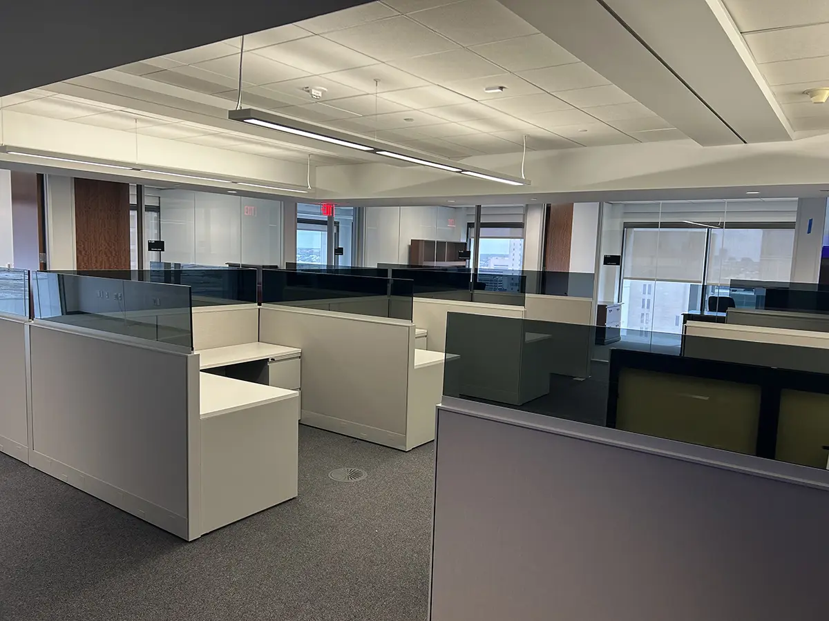 Modern office space with empty gray cubicles and glass partitions under fluorescent lights. Large windows reveal a cityscape in the background.