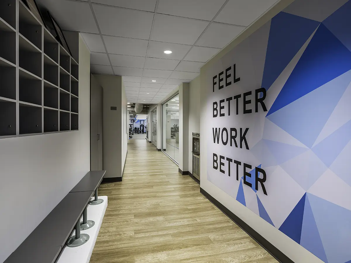A modern gym hallway with motivational text "Feel Better, Work Better" on a geometric blue-patterned wall, leading to a well-lit workout area.