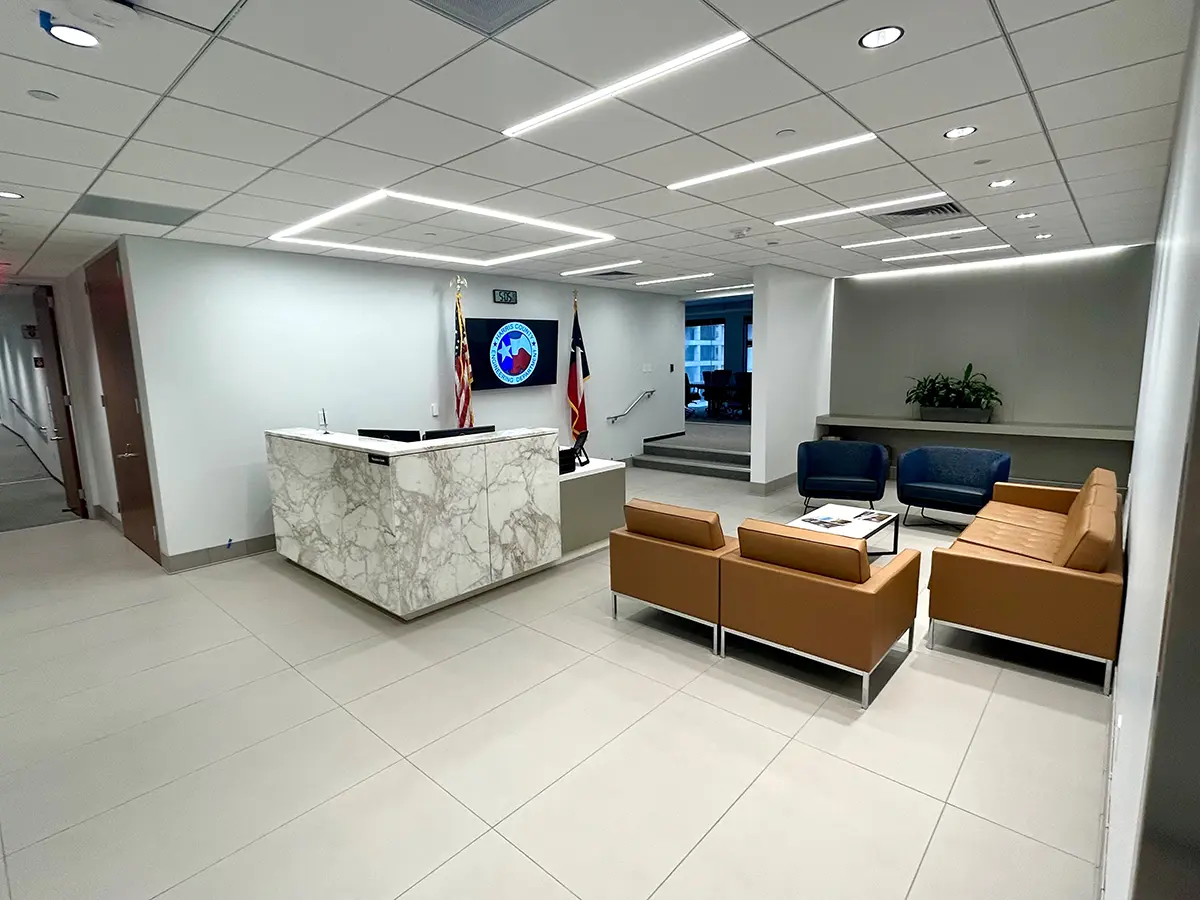 Modern lobby with a marble reception desk, wall flags, and a digital clock. Brown sofas and blue chairs offer seating. Bright lighting creates a welcoming atmosphere.