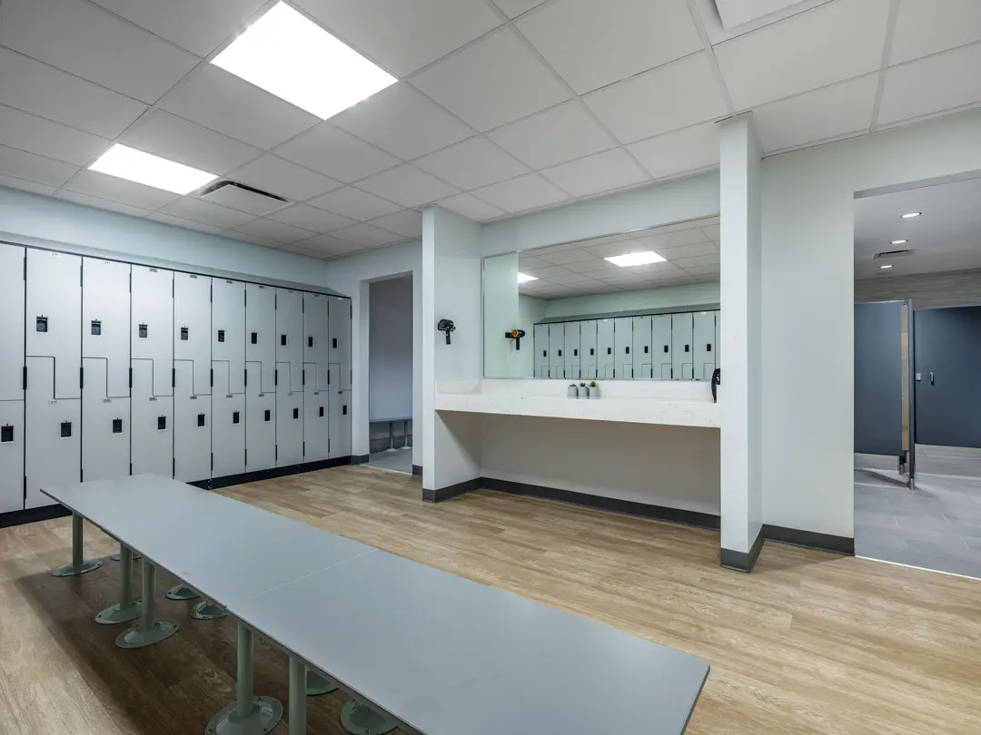 Bright locker room with a row of gray lockers on the left, a long bench in the center, and a wide mirror above sinks on the right. The tone is clean and modern.