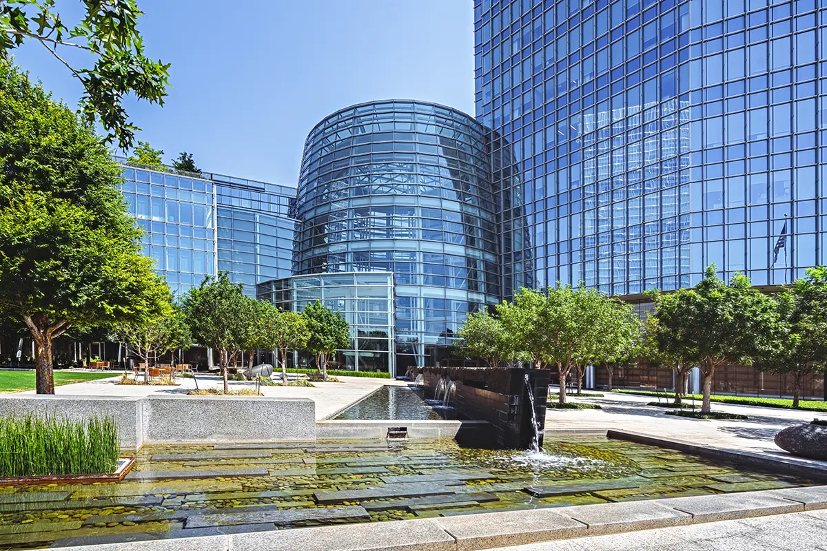 Modern glass buildings with a landscaped courtyard featuring a reflecting pool and trees. The scene is serene, with a clear blue sky overhead.