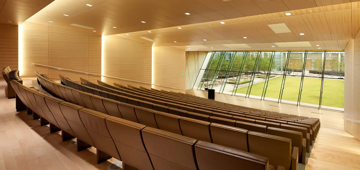 Modern auditorium with wooden walls and seating. Large glass windows offer a view of a green courtyard outside. Warm lighting creates a serene atmosphere.