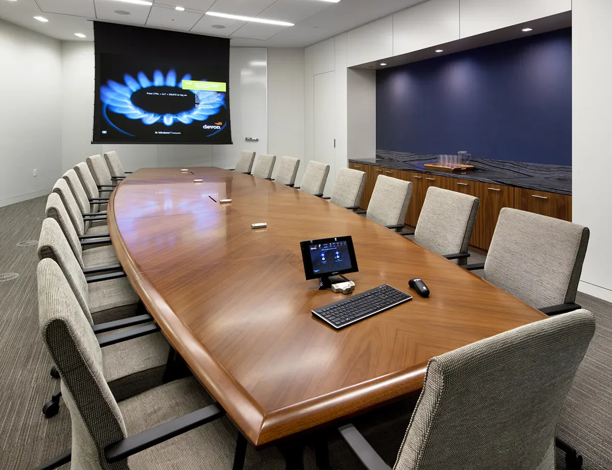 Spacious conference room with a long wooden table surrounded by beige chairs. A screen projects a blue graphic. Modern, professional atmosphere.