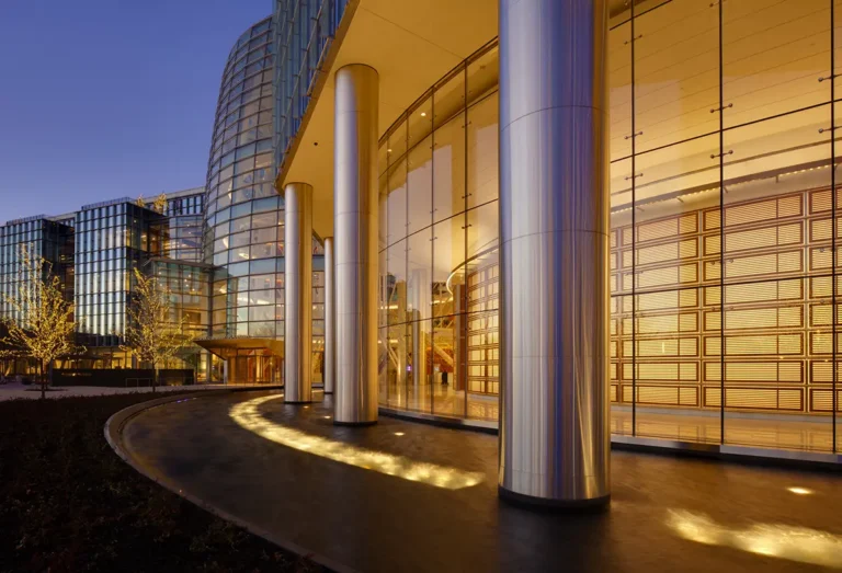 Modern glass building at dusk with tall, sleek aluminum columns and illuminated interior. Reflections and warm lights create an elegant, serene atmosphere.