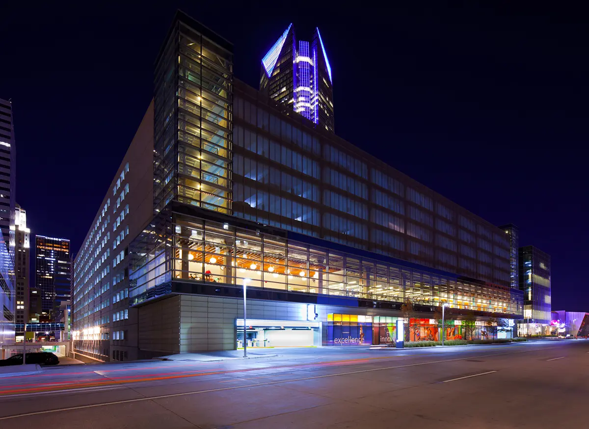 Nighttime image of an illuminated modern urban building with glass walls and a vibrant blue and purple rooftop glow, set against a dark sky.