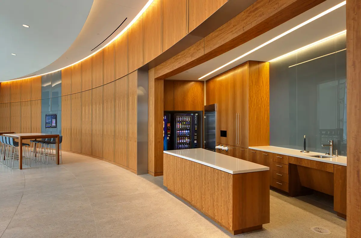 Modern kitchenette with wooden cabinets, a vending machine, and a white countertop. Curved wall with high stools and tables under soft lighting.
