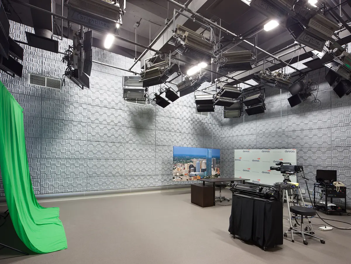 A spacious broadcast studio with a green screen to the left, overhead lighting, camera equipment, and a backdrop featuring a cityscape image and logos.