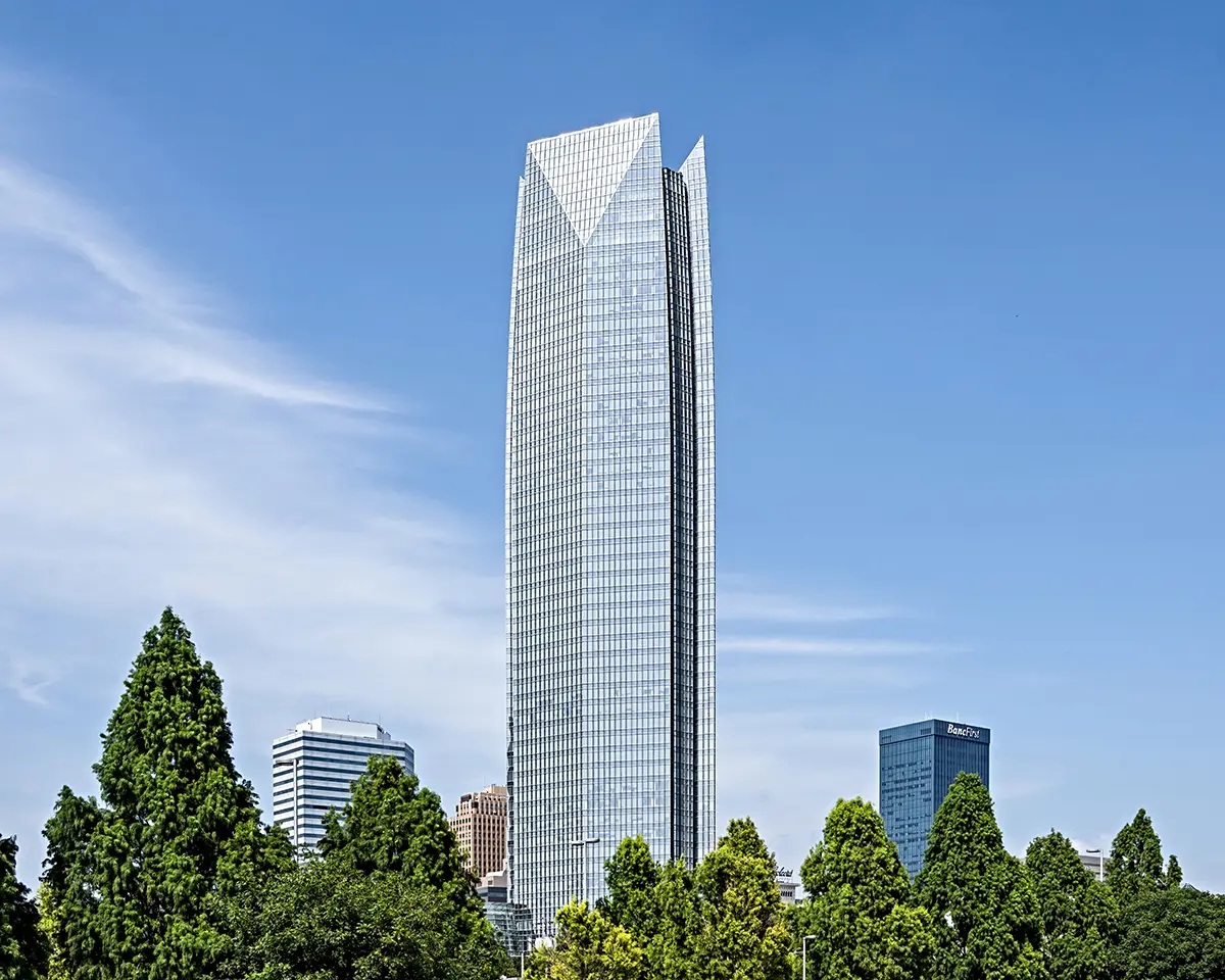 A tall, modern glass skyscraper rises against a clear blue sky, surrounded by lush green trees and smaller buildings. The scene is serene and impressive.