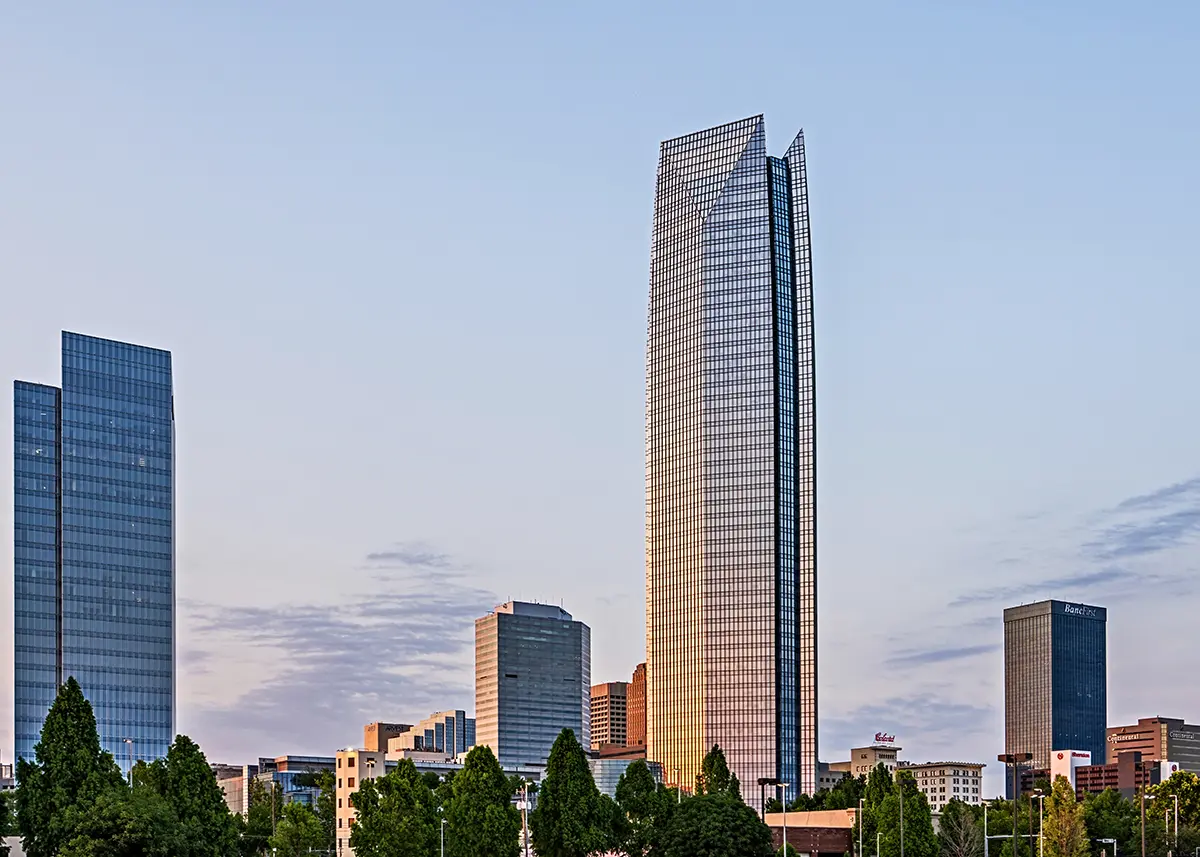 Cityscape at dusk with modern skyscrapers under a clear sky. Tallest building, center-right, reflects golden light. Green trees line the foreground.