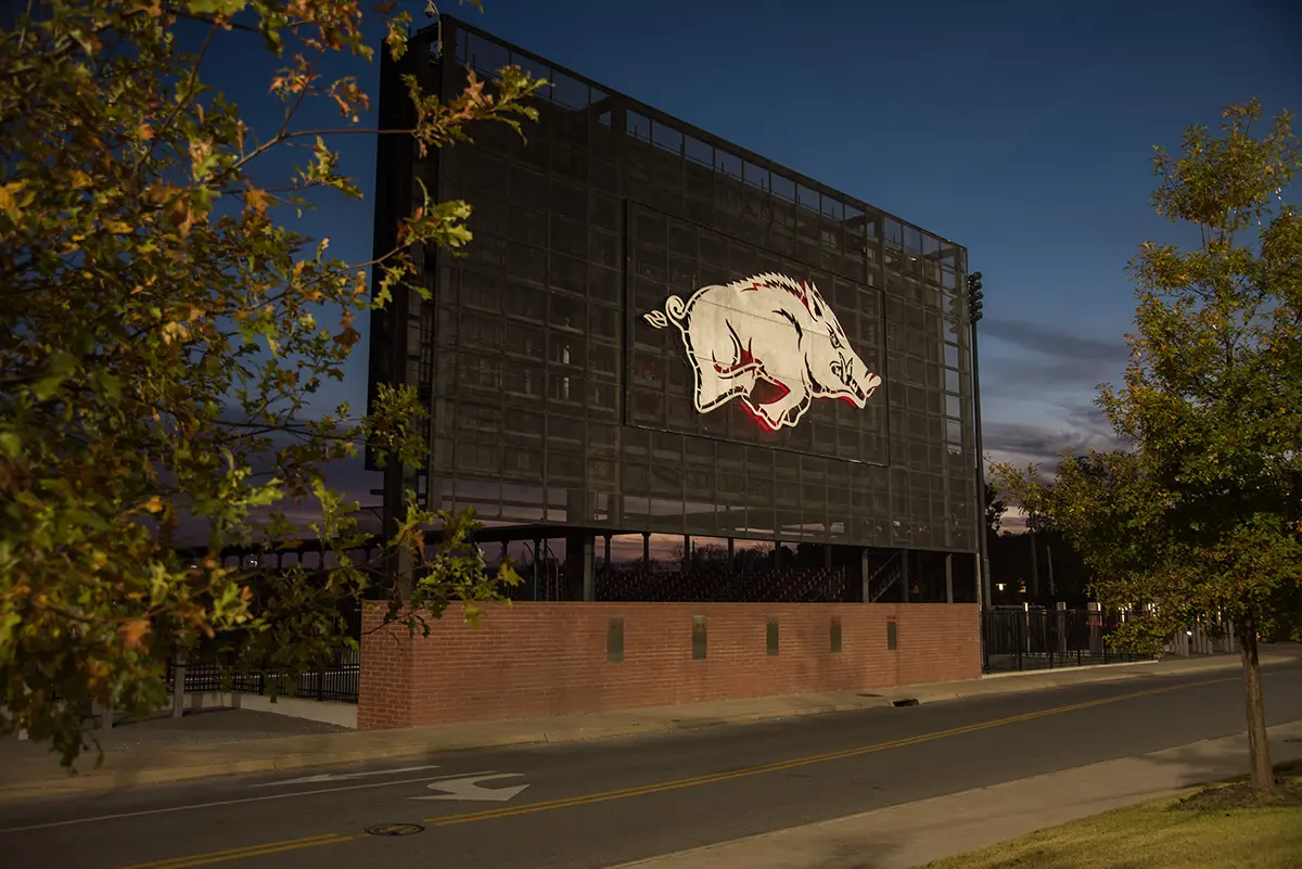 Large outdoor sign with a hog logo on a brick structure at dusk. Surrounded by trees and a road, the setting is calm with a blue and pink sky.