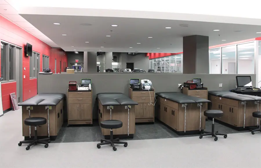 Athletic training room with five black exam tables, wooden cabinets, and stools. Medical equipment on counters. Red and white walls, bright lighting.