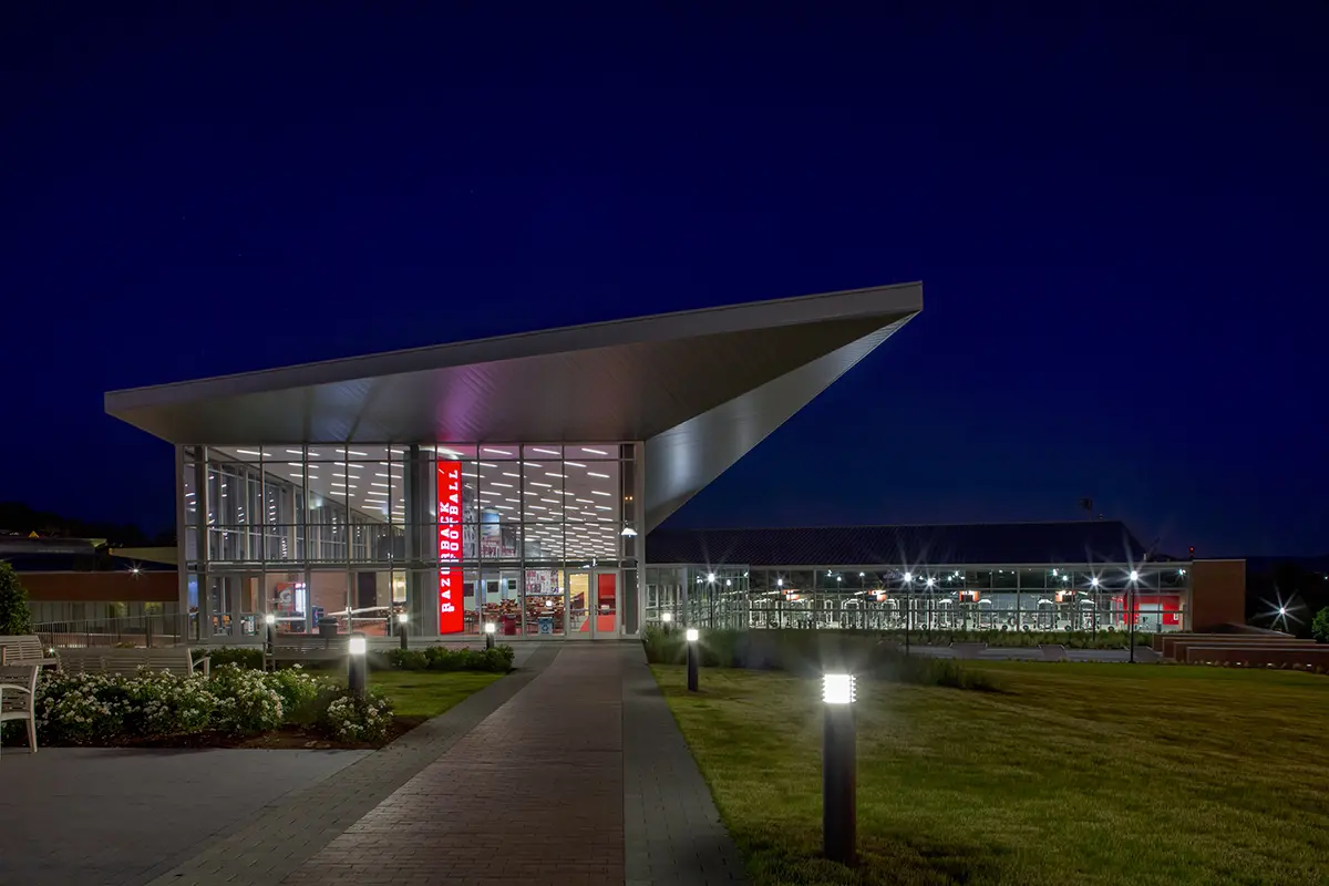 Modern lecture hall with tiered gray seating and red accents, featuring a large projection screen, red walls, and a boar logo on the carpet.
