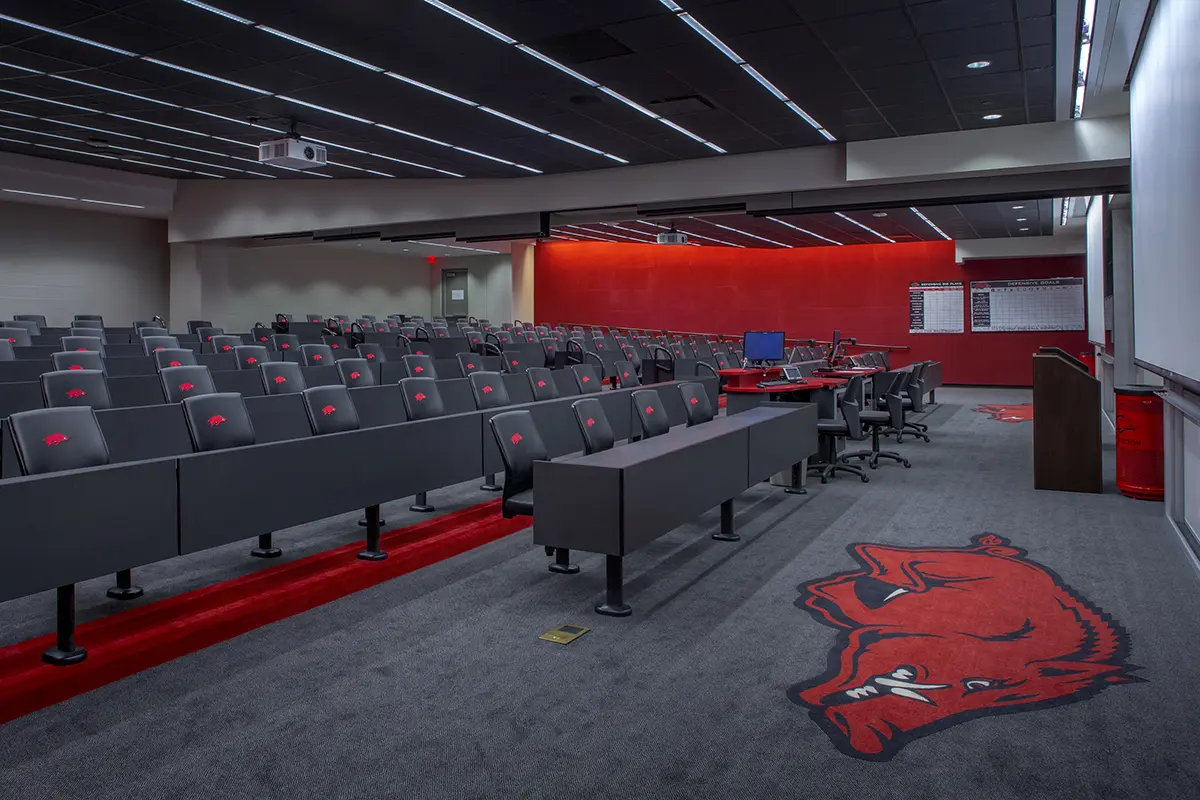 Modern lecture hall with tiered gray seating and red accents, featuring a large projection screen, red walls, and a boar logo on the carpet.