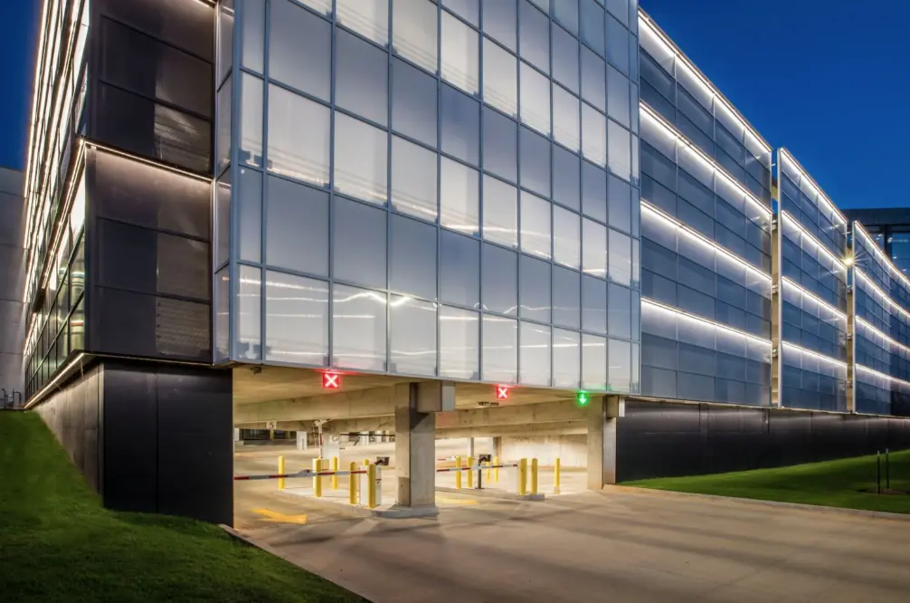 Modern parking garage at dusk, featuring illuminated glass panels and a clear entrance with red and green lights indicating availability.