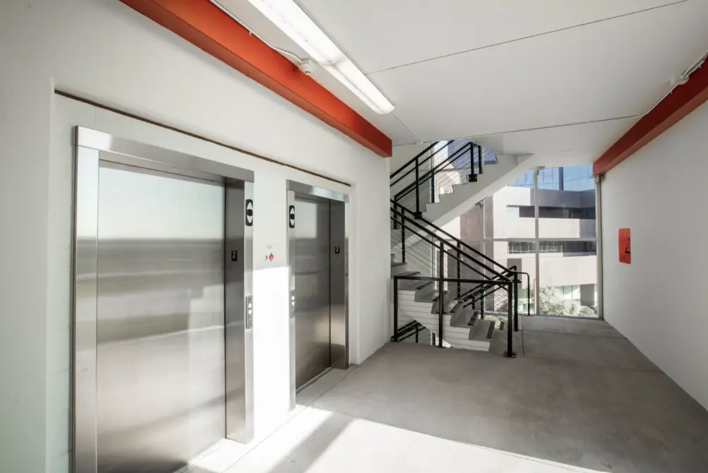 Modern, sunlit building hallway with two stainless steel elevators on the left and a staircase with metal railings by a large window on the right.