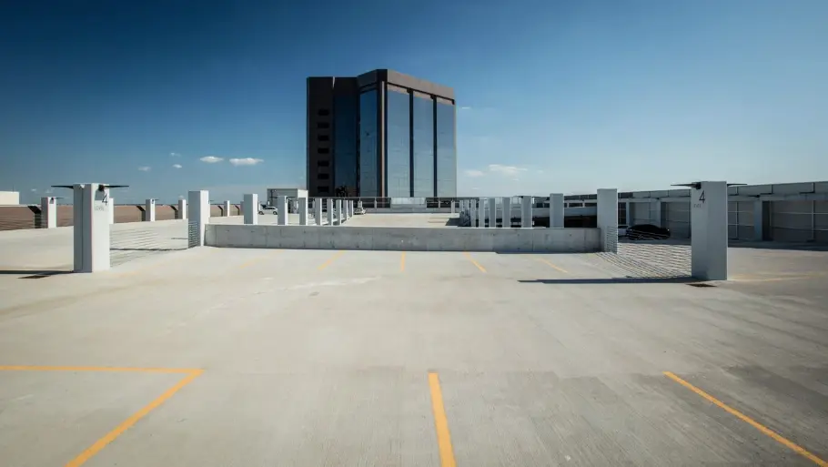 An empty rooftop parking lot under a clear blue sky, marked with yellow lines. A tall, modern glass building stands prominently in the background.