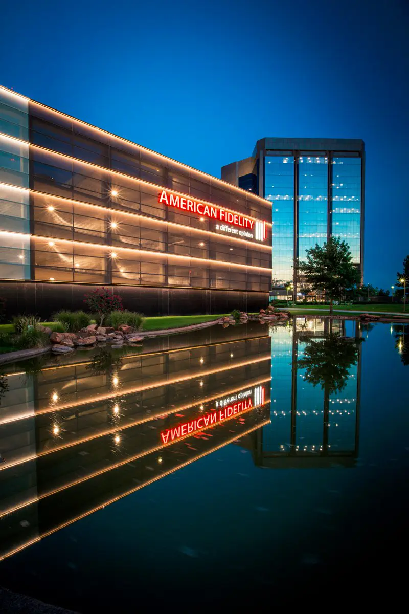 Modern office building with "American Fidelity" signage illuminated at night. Reflective glass facade and water create a serene, calm atmosphere.