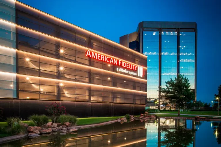 Modern office building with "American Fidelity" signage illuminated at night. Reflective glass facade and water create a serene, calm atmosphere.