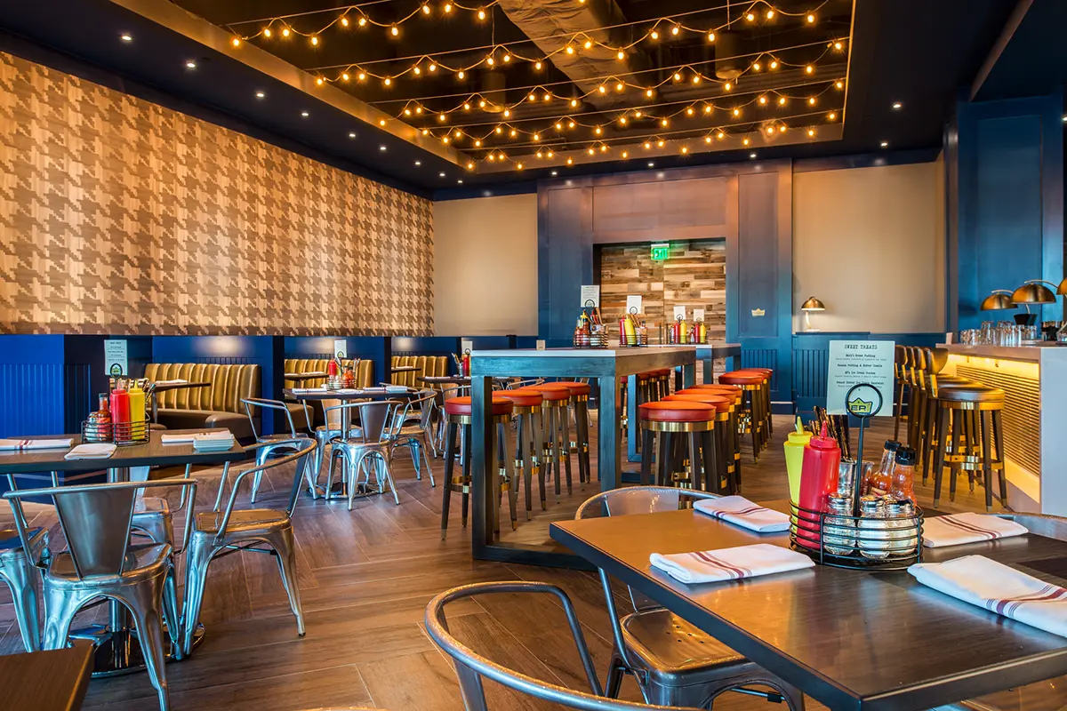 Modern restaurant interior with wooden floors, string lights on the ceiling, and tables set with napkins and condiments, creating a warm, inviting ambiance.