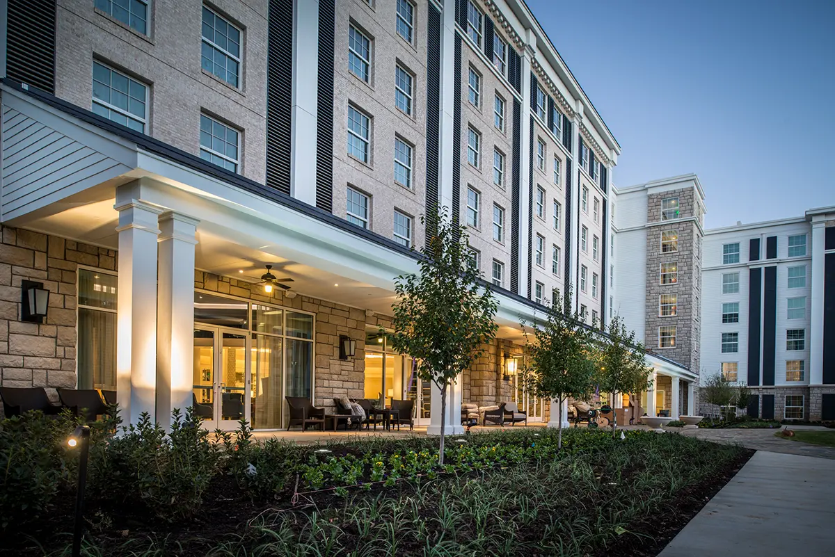 Modern hotel exterior at dusk, showcasing a multi-story building with illuminated windows, a covered patio, and landscaped gardens creating a welcoming atmosphere.