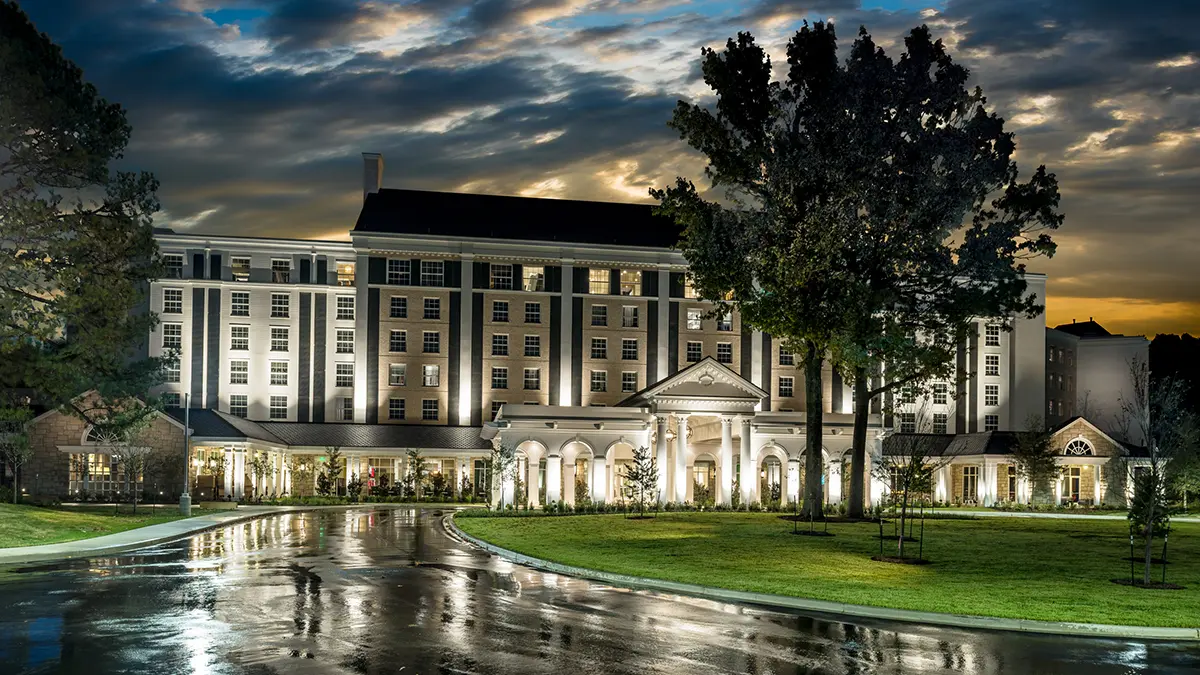 Illuminated grand hotel at dusk, with dramatic clouds overhead. Reflective wet driveway leads to the entrance, surrounded by trees and manicured lawns.