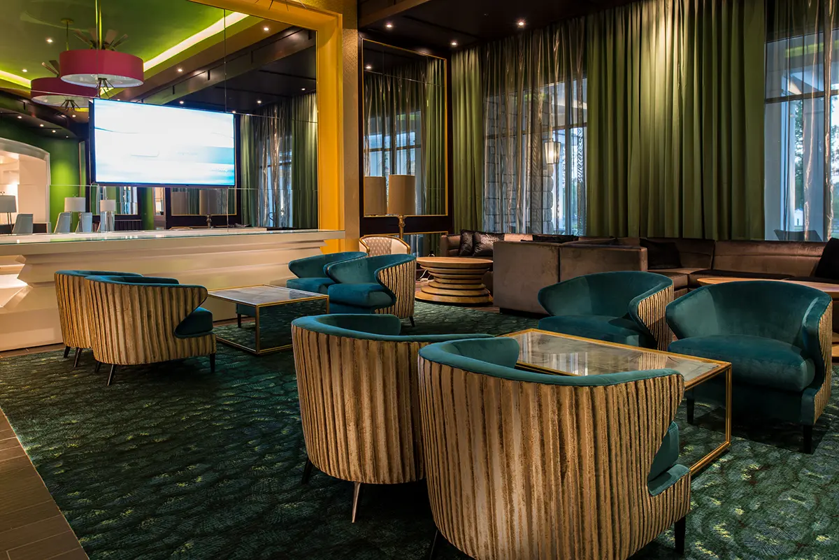 Stylish hotel lobby with teal velvet chairs, striped armchairs, and glass tables on green carpet. Green curtains and a large screen create a cozy ambiance.