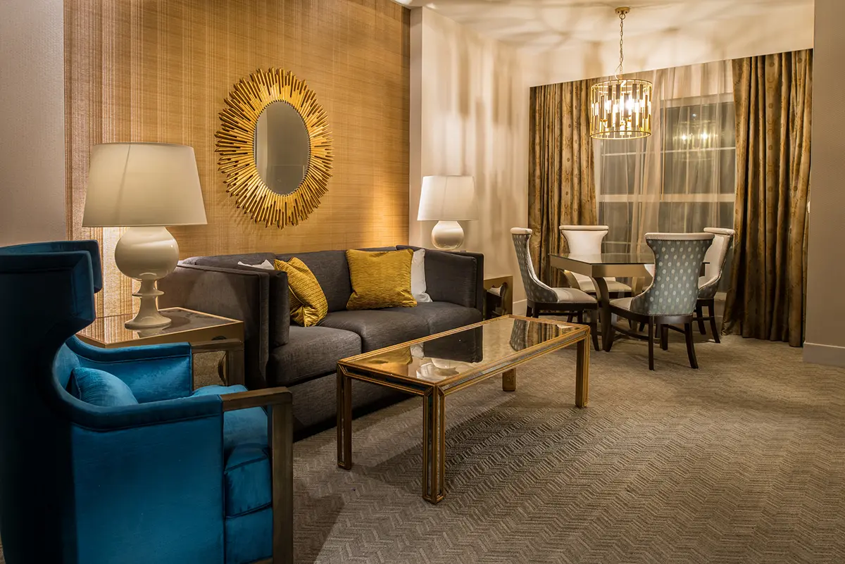 Elegant living room with a gray sofa, blue armchair, and gold accents. Sunburst mirror on a textured wall, and a round dining table with modern chairs. Warm lighting.