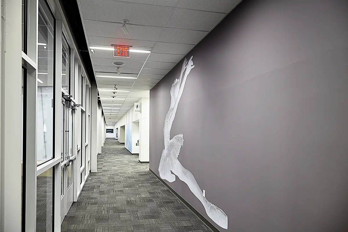 A sleek hallway with gray carpet and glass doors features a large, graceful dancer mural on the right wall, adding elegance and artistic flair.