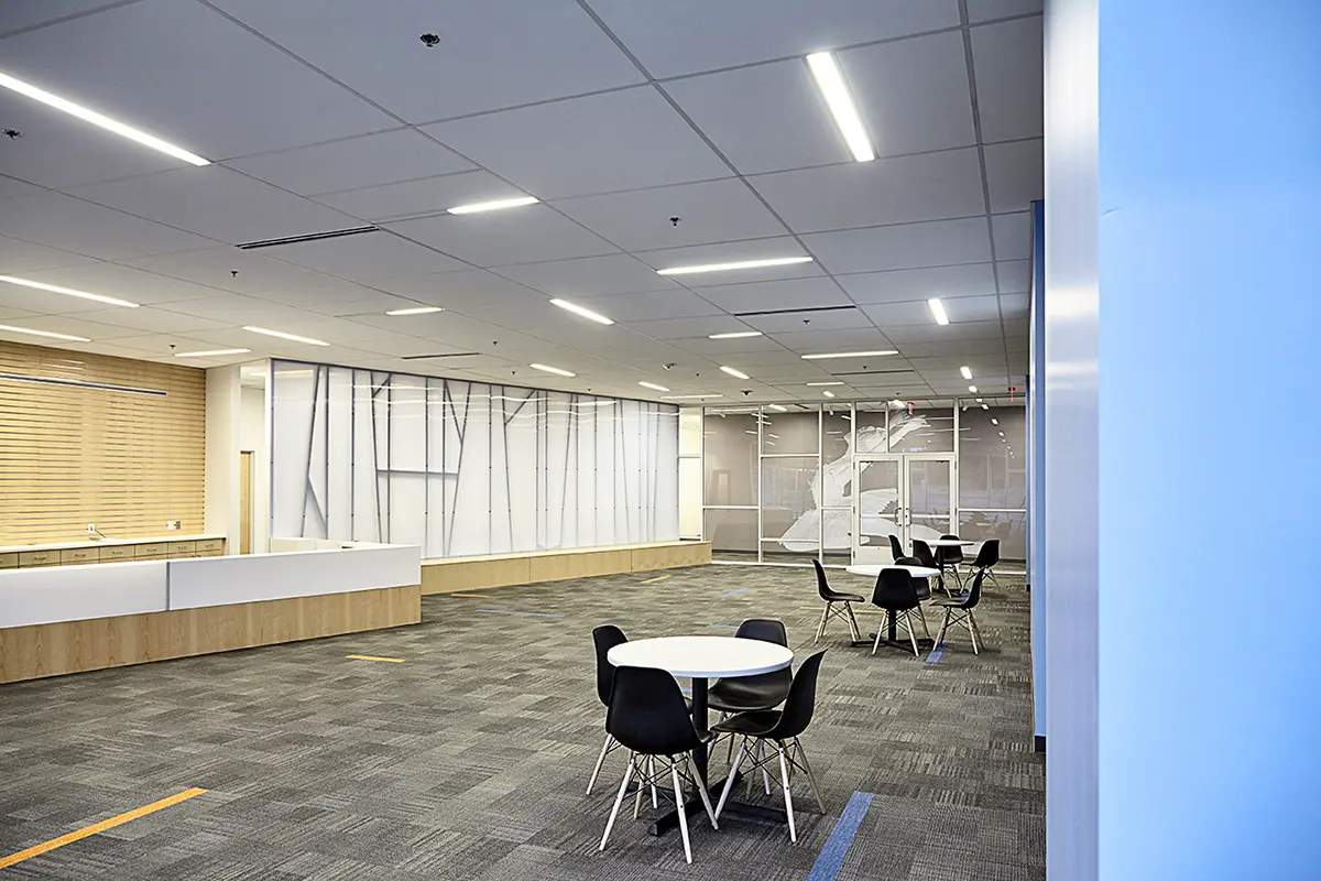 Spacious modern office with a minimalist design, featuring round tables with black chairs, a textured wall, and a ceiling of linear lights.
