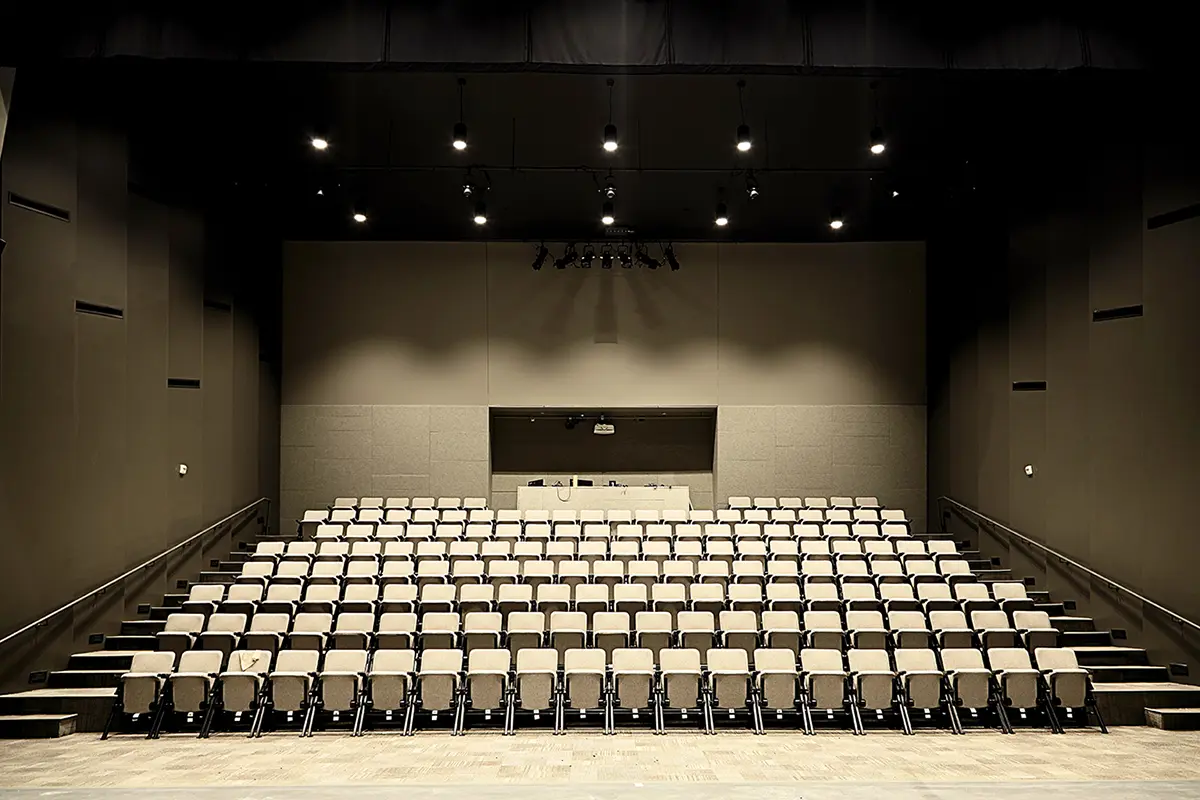 An empty theater with rows of beige seats facing a stage. The dim lighting and minimalist design create a quiet and anticipatory atmosphere.