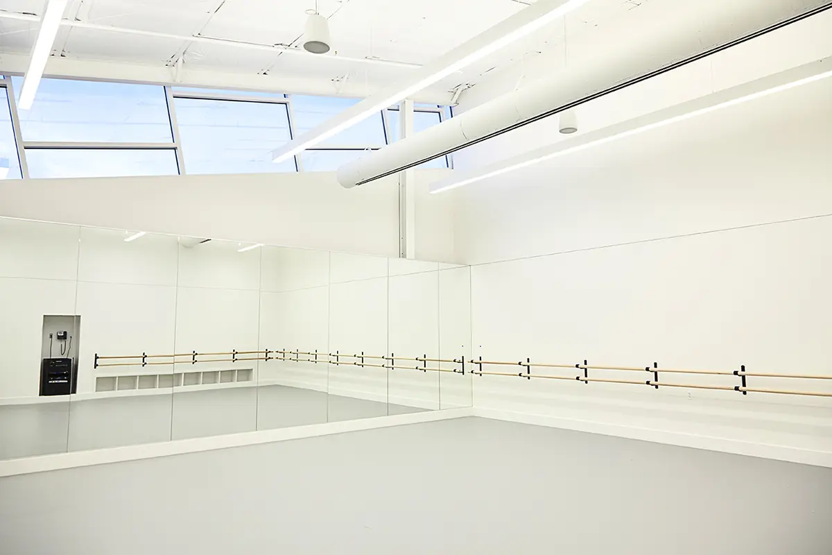 A bright, empty dance studio with large mirrors, wooden ballet barres, and high ceiling windows. The space feels spacious, modern, and inviting.