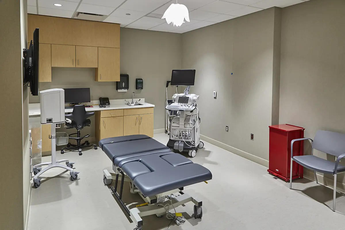 Una moderna sala de exploración médica con una camilla ajustable de color gris oscuro, un ecógrafo, armarios de madera, un escritorio con ordenador y sillas grises, que transmite un ambiente clínico.