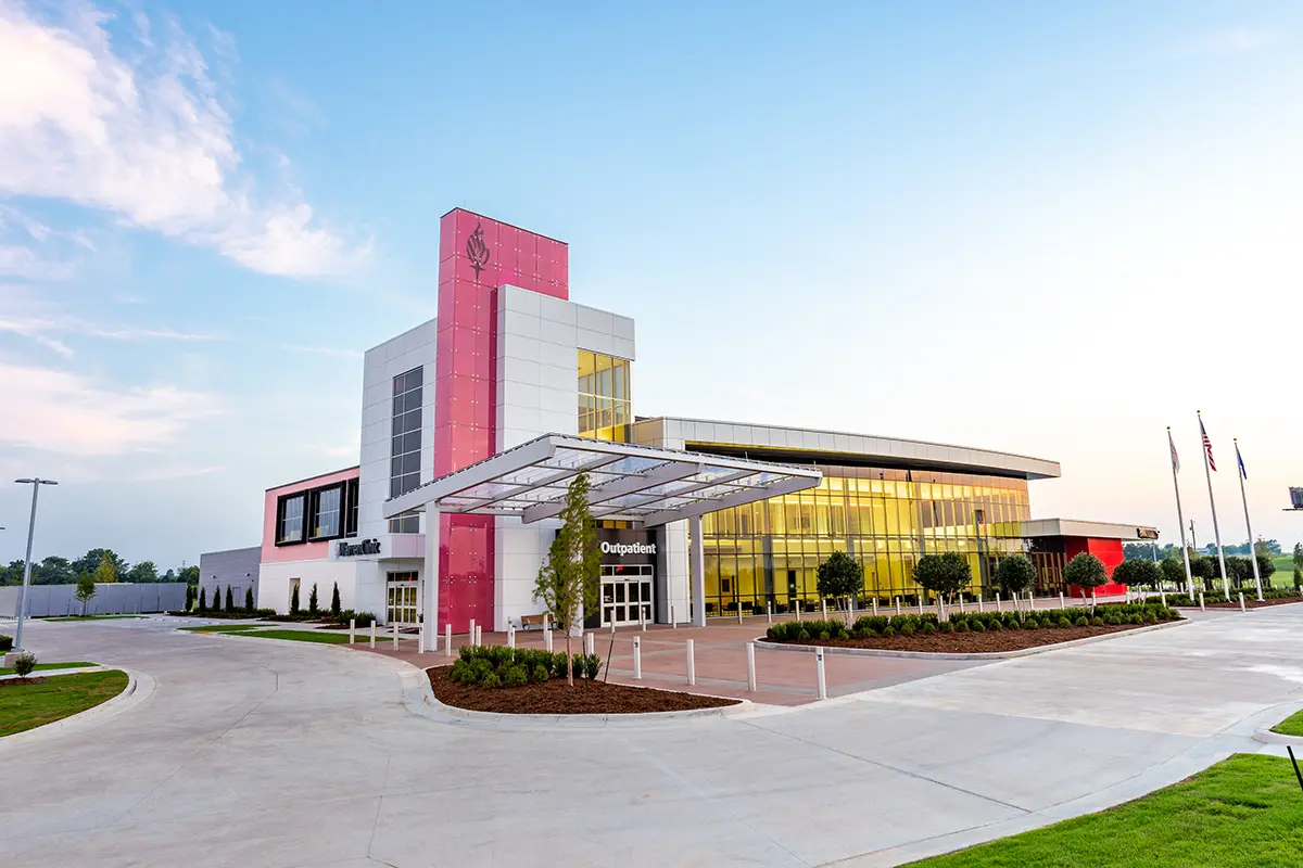 Exterior de un moderno centro médico con una llamativa fachada rosa y blanca, grandes ventanas de cristal que reflejan el cielo, rodeado de cuidados jardines y banderas.