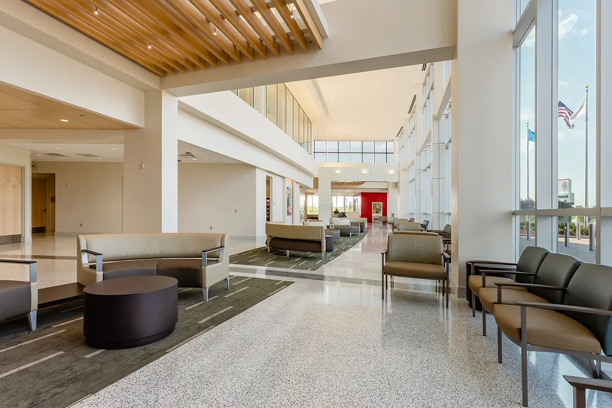 Amplio vestíbulo con modernos asientos, detalles en madera en el techo, grandes ventanas y banderas en el exterior. Ambiente luminoso y acogedor con luz natural.