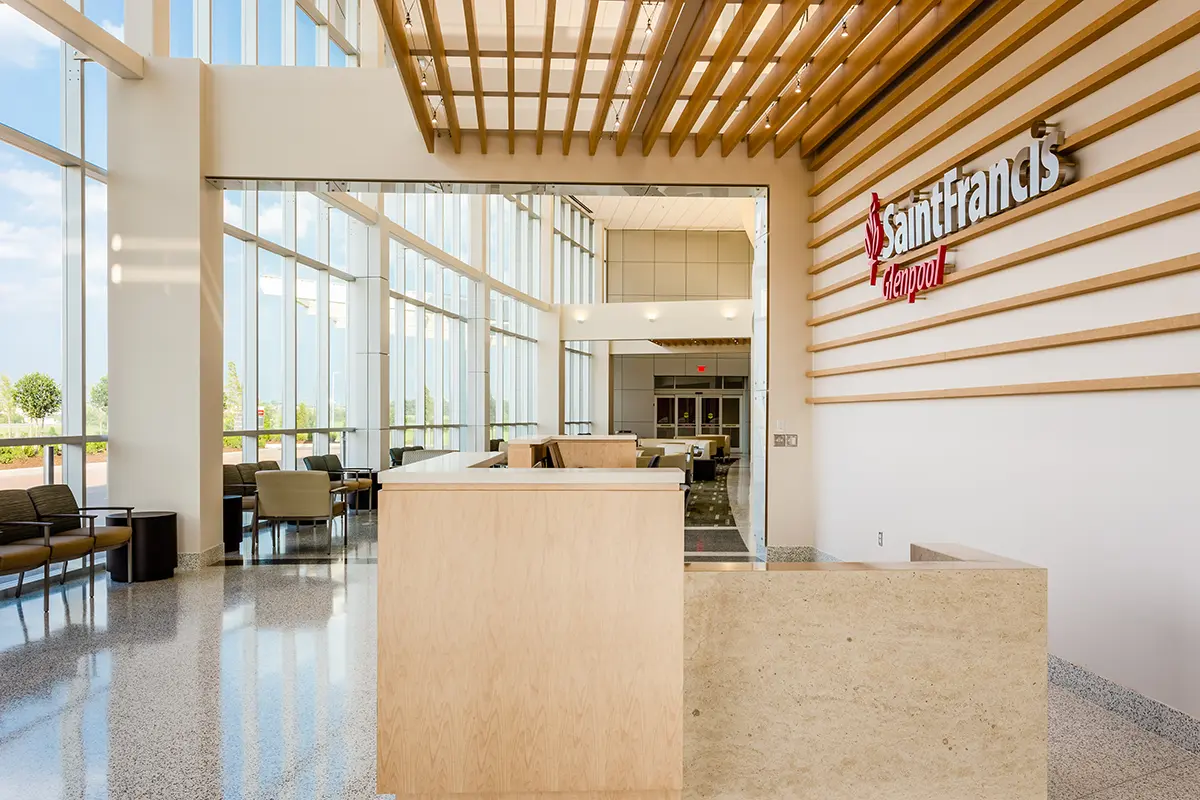 Luminoso vestíbulo del hospital con techos altos y grandes ventanas. Los detalles en madera clara y el mobiliario moderno crean un ambiente acogedor y espacioso. El letrero dice «Saint Francis».