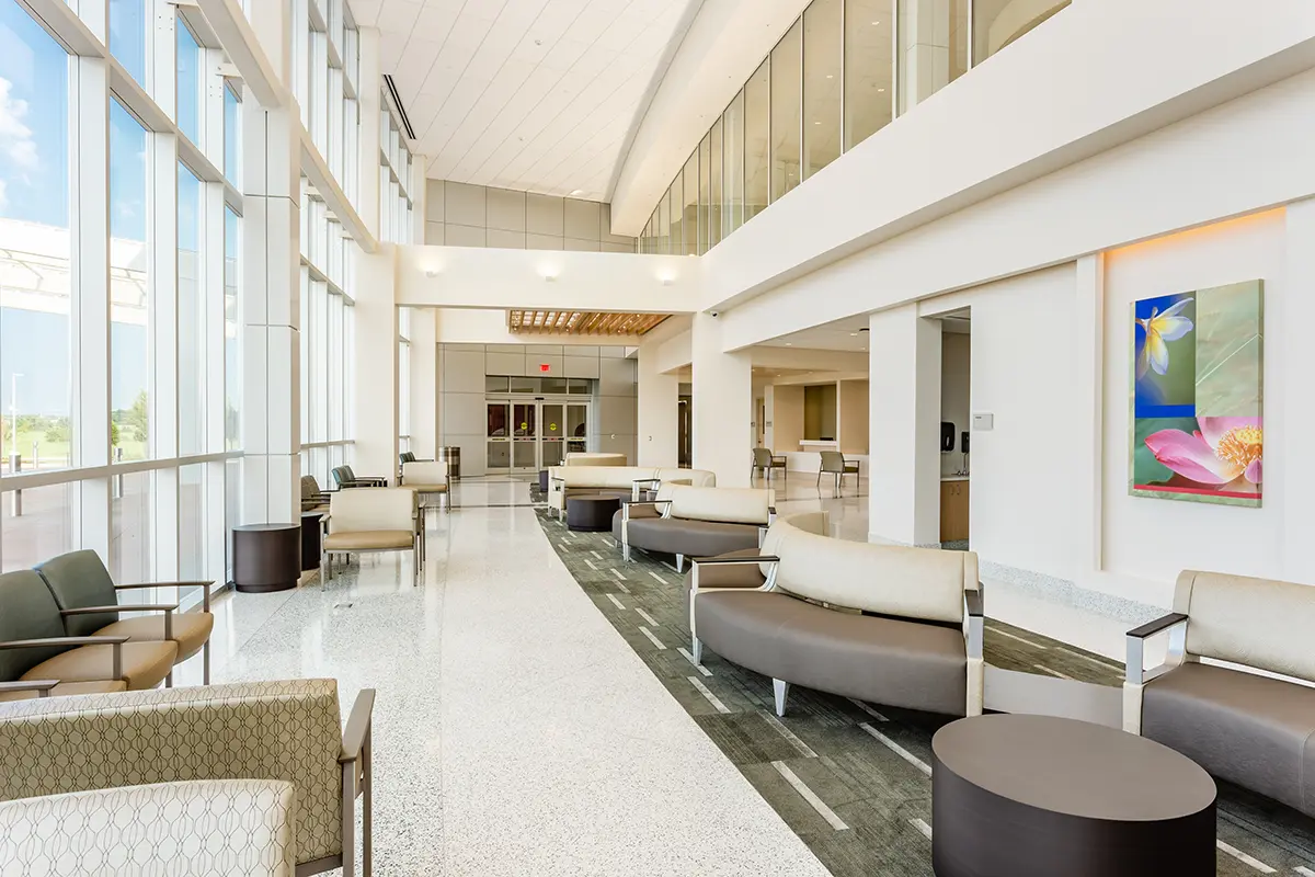 Amplio y moderno vestíbulo con grandes ventanales, luz natural y elegantes asientos. Una vibrante obra de arte floral adorna la pared, creando un ambiente acogedor.