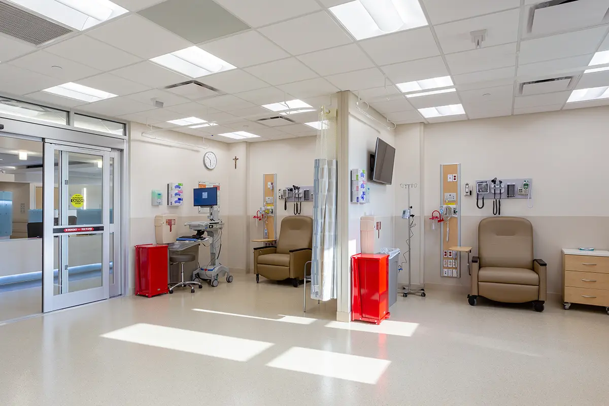 Una habitación de hospital limpia y bien iluminada con dos sillones reclinables, equipo médico, contenedores de almacenamiento rojos y un televisor montado en la pared. El ambiente es estéril y ordenado.