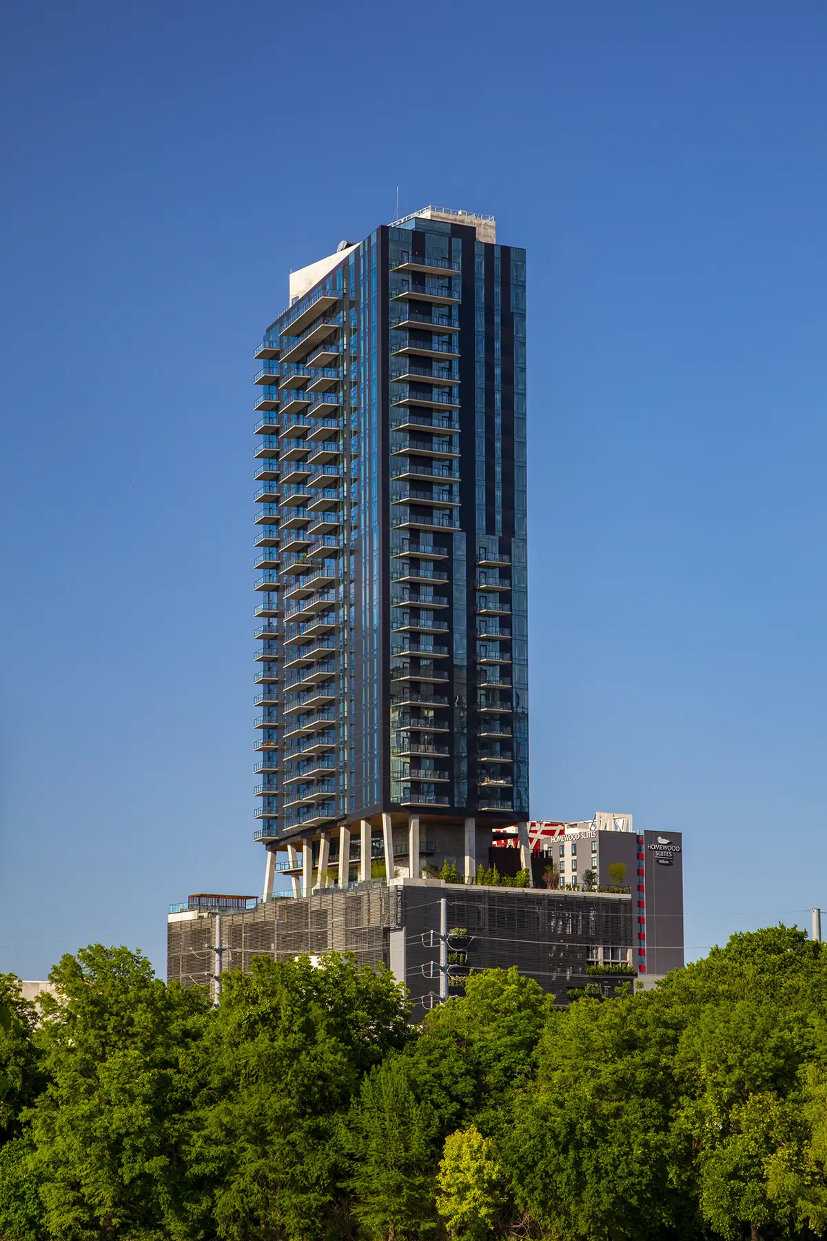 Tall modern skyscraper with glass facade and balconies set against a clear blue sky. Lush green trees in the foreground create a vibrant contrast.