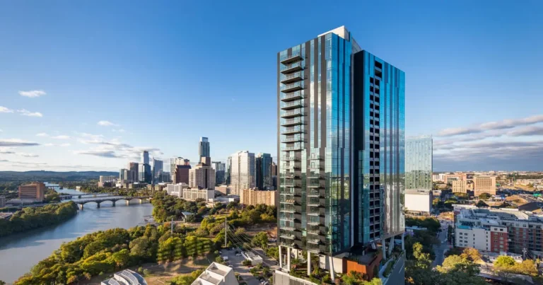 A tall, modern glass skyscraper stands prominently in a city skyline with a river to the left. The setting is bright and clear, evoking a sense of vibrancy.