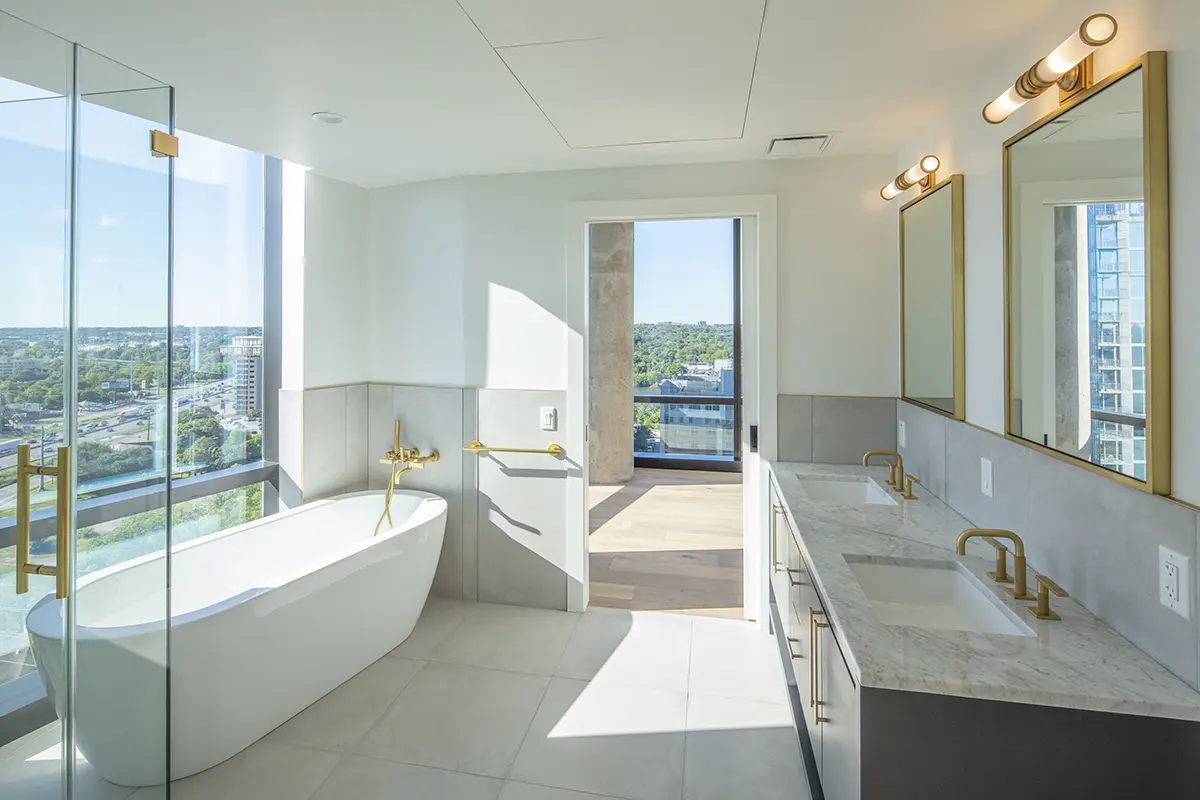 Luxurious bathroom with large windows offering a scenic city view. Features a freestanding tub, glass shower, dual sinks with gold fixtures, and bright, natural light.