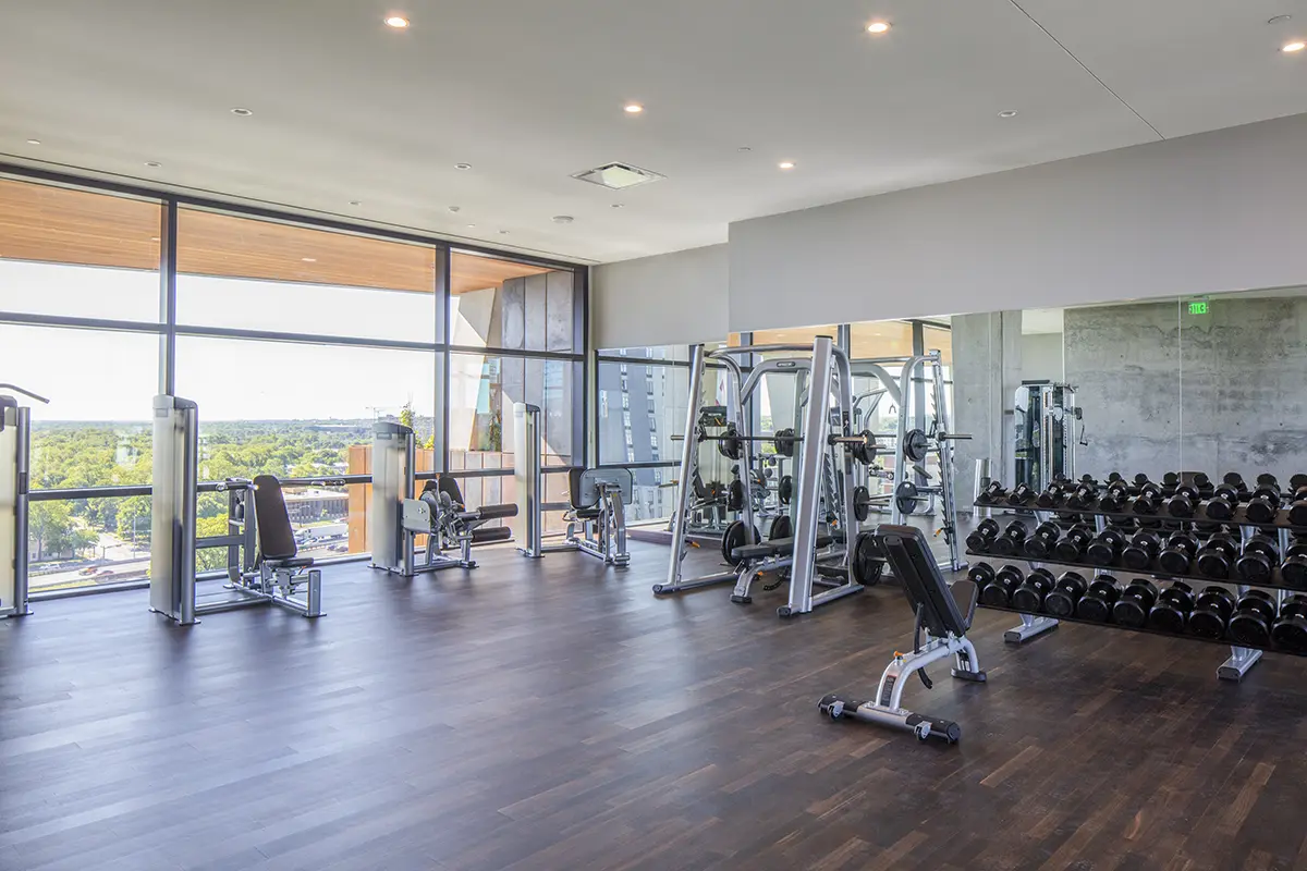 Bright, modern gym with large windows and a city view. Features weight machines, dumbbells, and sleek dark wood flooring, creating a clean, airy atmosphere.