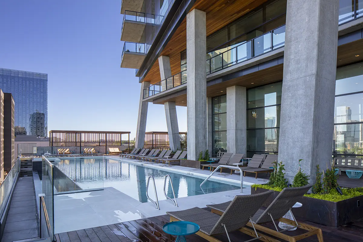 Rooftop pool with clear blue water, surrounded by modern lounge chairs and greenery. Overlooks city skyline under a clear blue sky, creating a serene atmosphere.