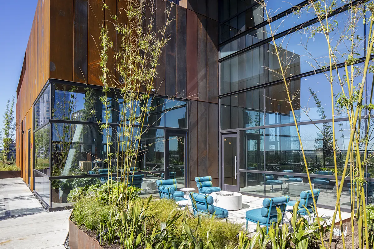 Modern building exterior with glass walls and rust-colored panels. Outdoor seating area with turquoise chairs and white tables amidst lush plants.