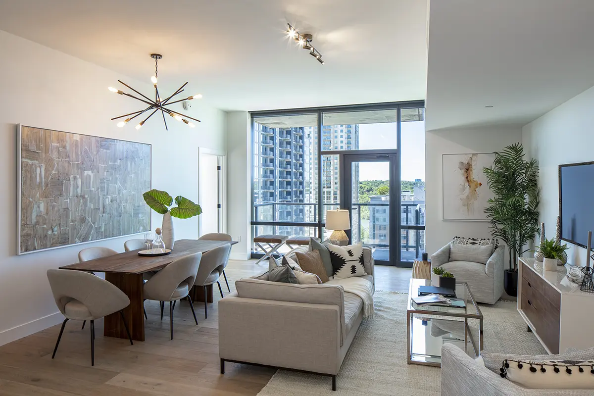 Modern living and dining area with neutral tones, featuring a stylish chandelier, large abstract art, cozy sofas, a wooden table, and cityscape views.