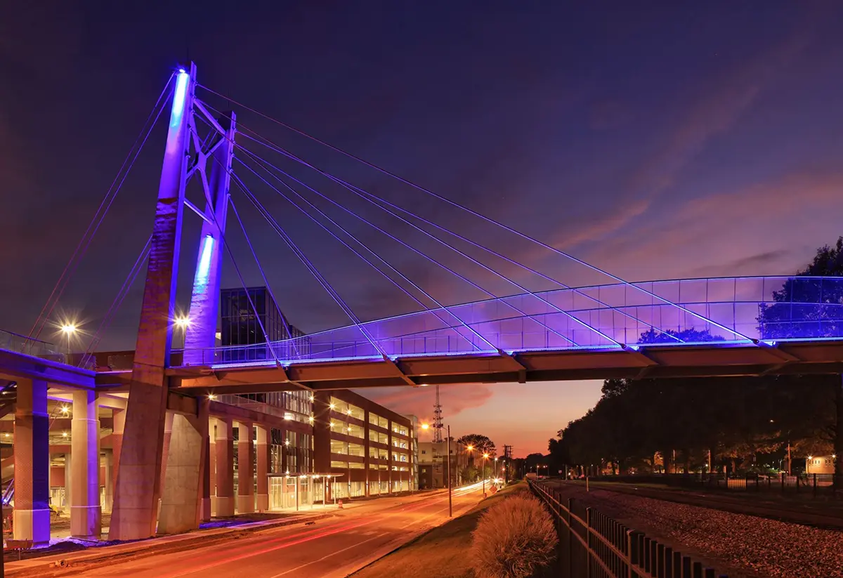 A modern cable-stayed bridge with vibrant blue lights spans an urban street at dusk. The scene conveys a futuristic and serene atmosphere.