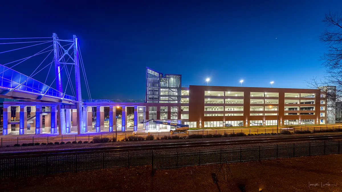 A modern, multi-level parking garage illuminated at night with blue lights highlighting its structure. The sky is deep blue, creating a calm, urban scene.