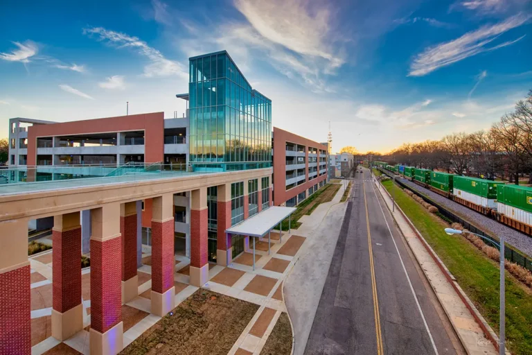 Modern building with glass facade and red brick pillars beside a road and green trains. Sky is partly cloudy at sunset, conveying a serene atmosphere.