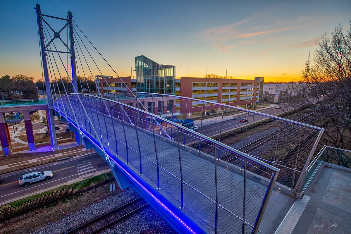 A modern pedestrian bridge with blue lights stretches over train tracks. A sunrise sky glows in the background, with urban buildings lining the street below.